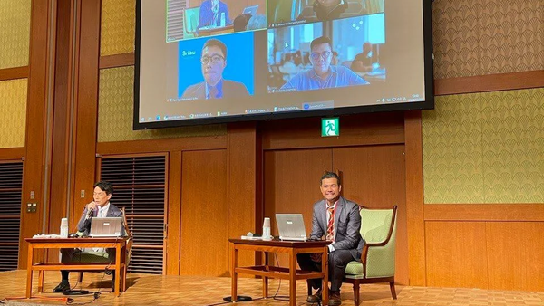 Two men sitting at tables on a stage with laptops, facing a large screen showing a four-person video call.