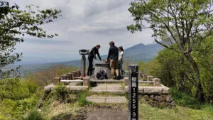 Photograph of hikers at a mountain summit marker with a sign reading "標高1255.9米" overlooking a valley in Nagano.