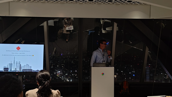 Photograph of Ben Allen speaking at a Tokyo Python meetup presenting "Building and Deploying Monorepo Workspaces" against a city skyline backdrop.